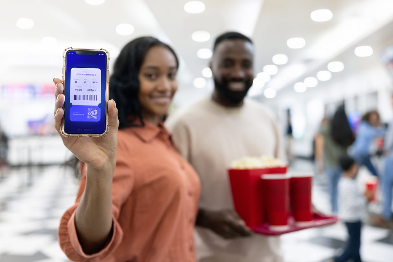 A woman holds up a smartphone displaying movie tickets, standing beside a man with popcorn and drinks at a brightly lit cinema lobby—showcasing smart movie theater hacks for a hassle-free night out.
