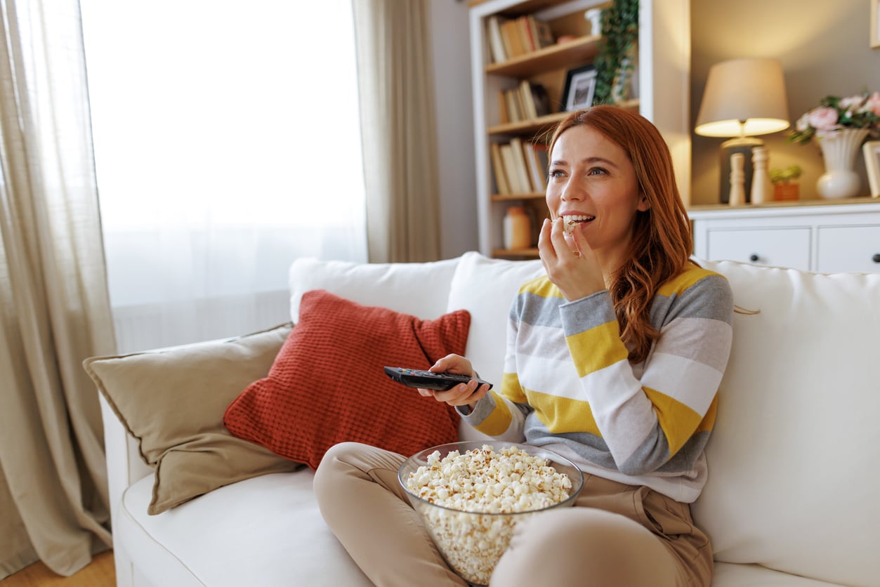 A woman sits on a couch eating popcorn and holding a remote, smiling while watching TV in a cozy living room with warm lighting and decorative pillows, embodying how women are changing their spending behaviors by finding joy in simple, comforting experiences at home.