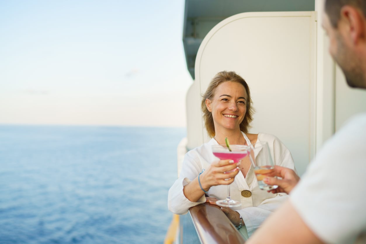 A woman smiles and holds a pink cocktail while clinking glasses with someone on a ship’s balcony overlooking the ocean, savoring the moment without worrying about vacation costs.