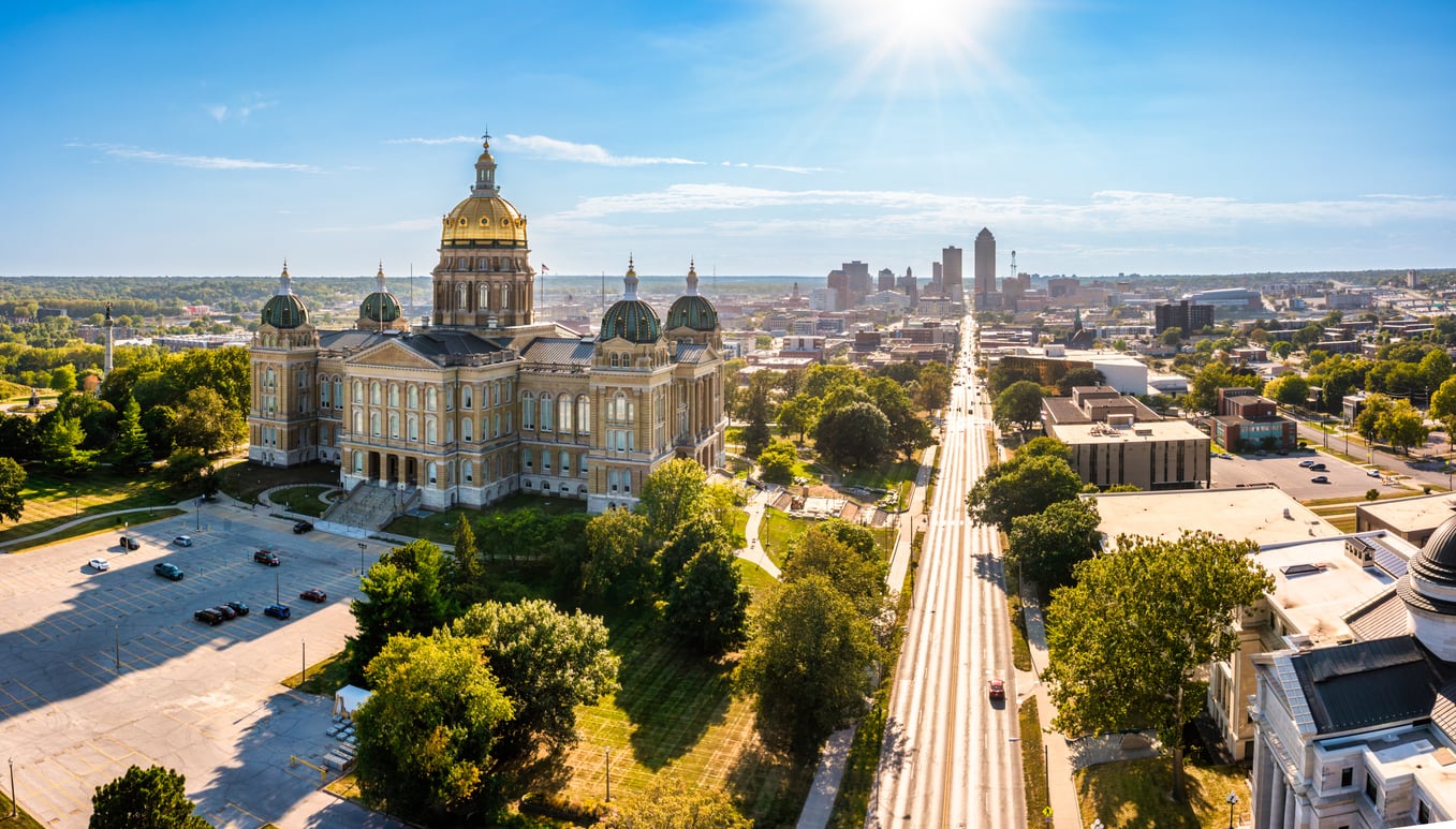 Aerial view of a large domed capitol building surrounded by trees, with a city skyline and sunlit sky in the background on a clear, travel-friendly day.
