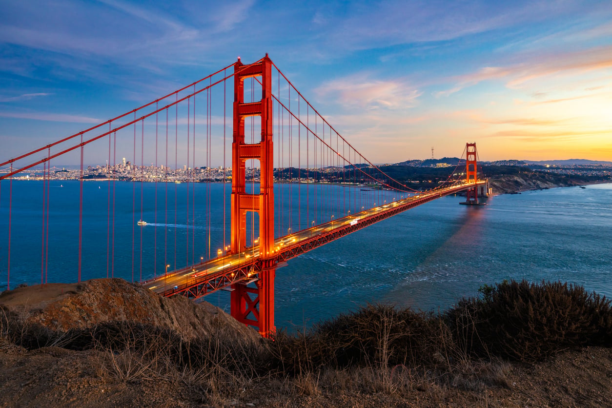 Golden Gate Bridge at sunset with lights on, spanning across blue water toward San Francisco city skyline, viewed from a hillside with some dry bushes in the foreground. The sky is partly cloudy with warm and cool tones.