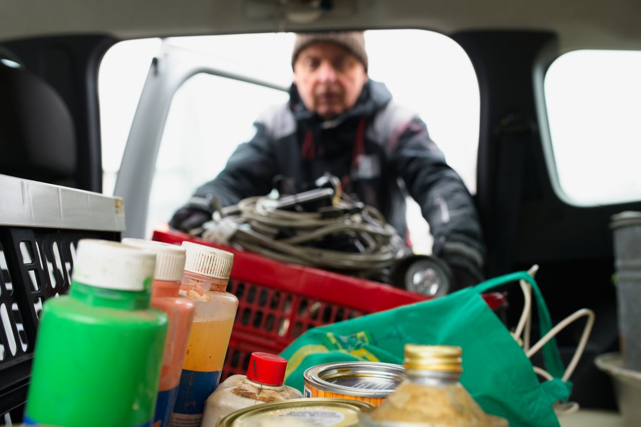 A person loads various supplies, including paint bottles, cans, and a plastic basket—perhaps even some car maintenance hacks—into the back of a vehicle. The scene is viewed from inside the car, with the person slightly out of focus in the background.