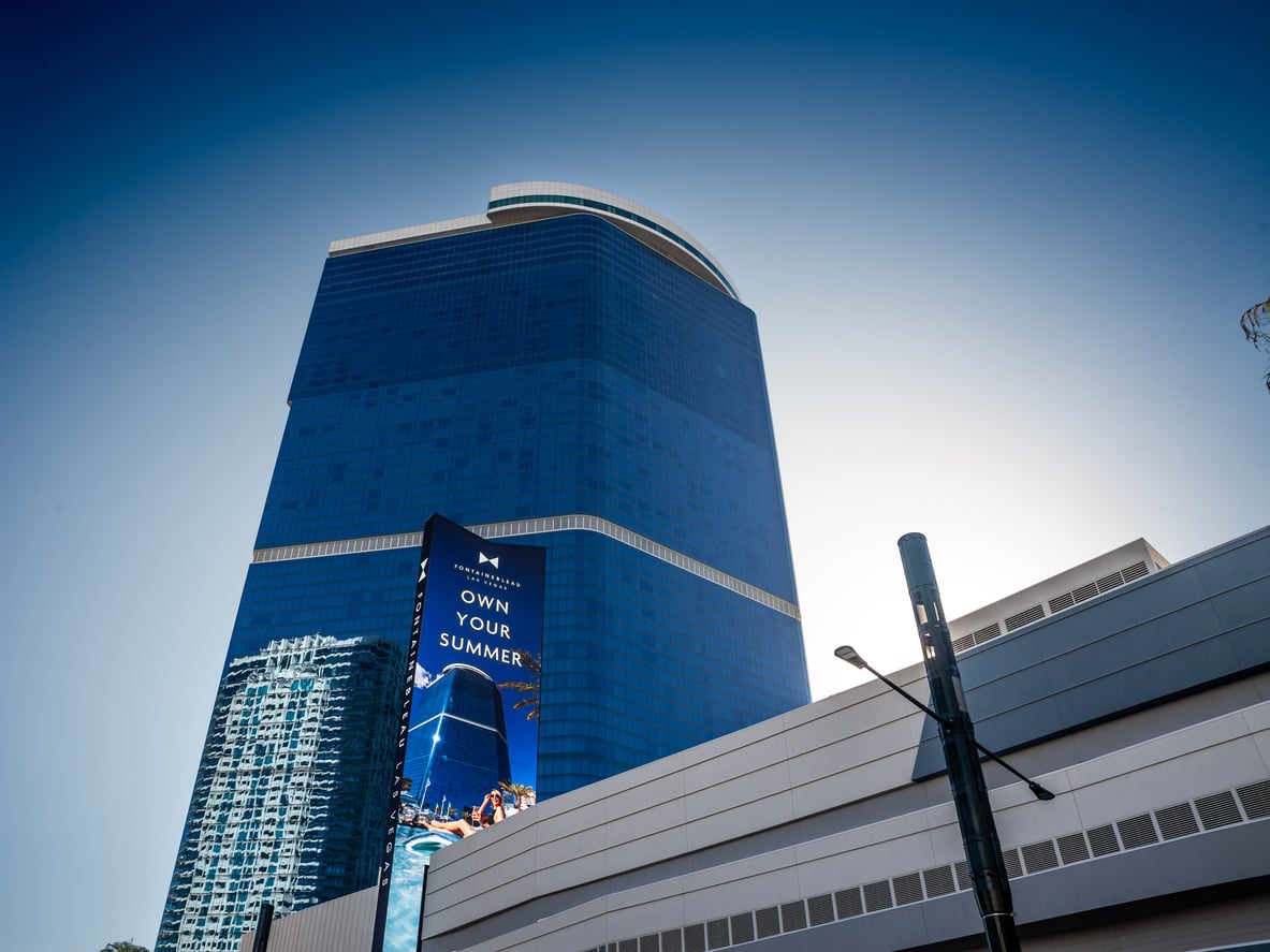 A tall blue glass skyscraper with a curved top is seen against a clear sky. A digital billboard on the building displays “OWN YOUR SUMMER.” Part of a white structure and another building are visible in front.