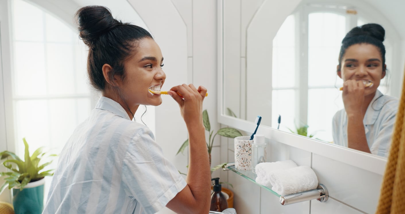 A woman with dark hair in a bun smiles while brushing her teeth in a bright bathroom. She stands by a sink with a towel, plants, and toothbrush holder visible. Her reflection shows in the mirror.