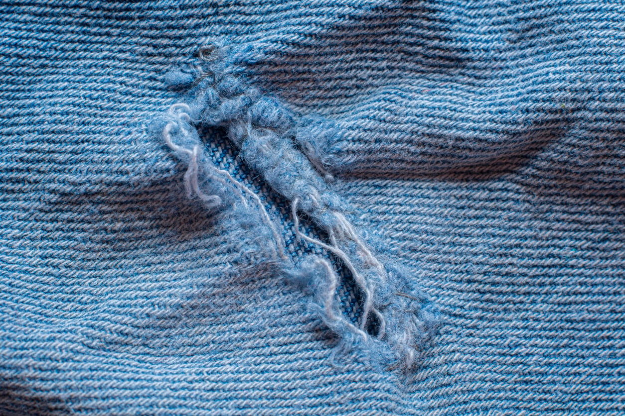 Close-up of a tear in light blue denim fabric, with frayed threads and rough edges surrounding the hole—a reminder of products that don't last.