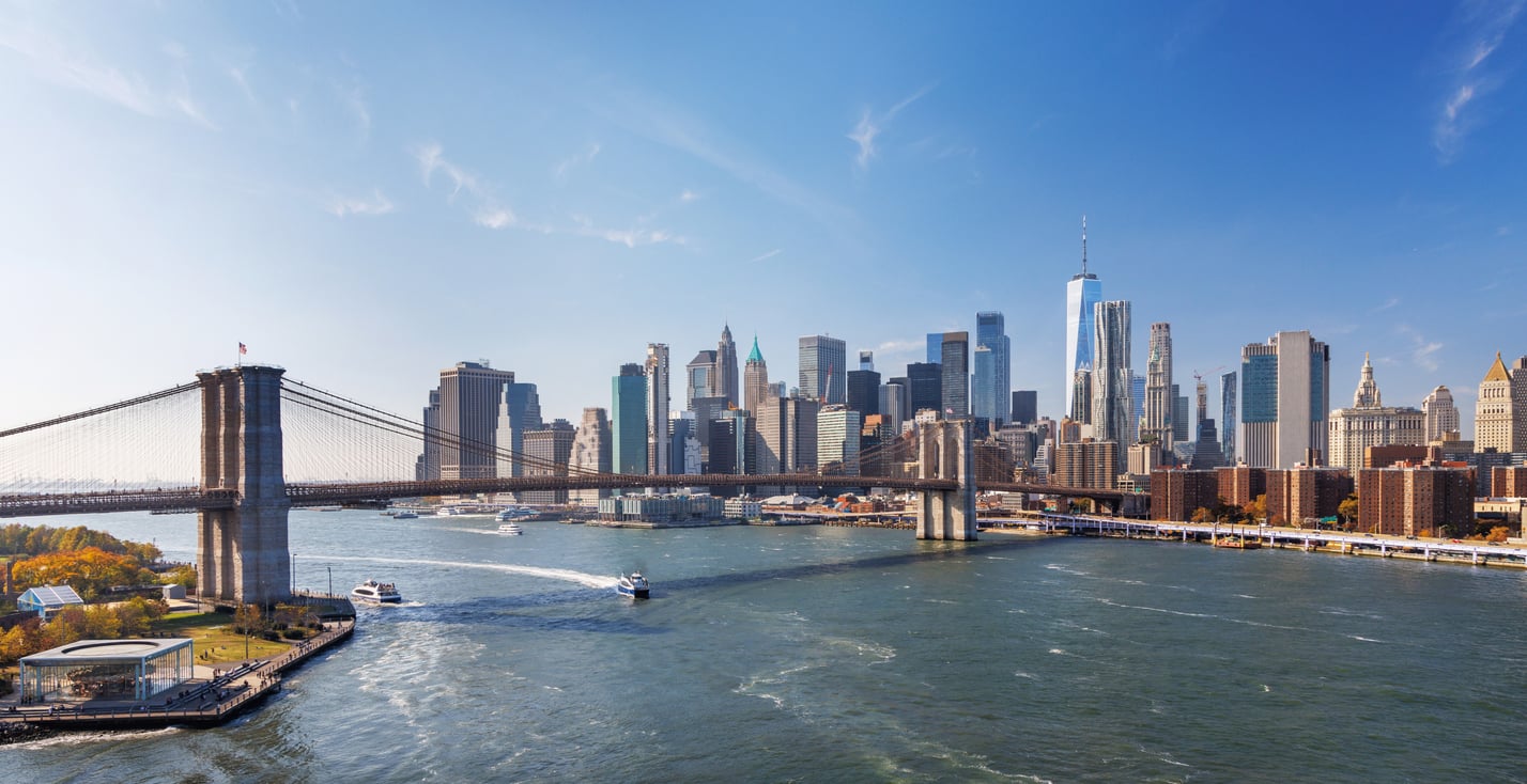 The iconic Brooklyn Bridge spans the East River with boats passing below, while the Manhattan skyline—including One World Trade Center—rises in the background under a clear blue sky, epitomizing the vibrant spirit of the East Coast.