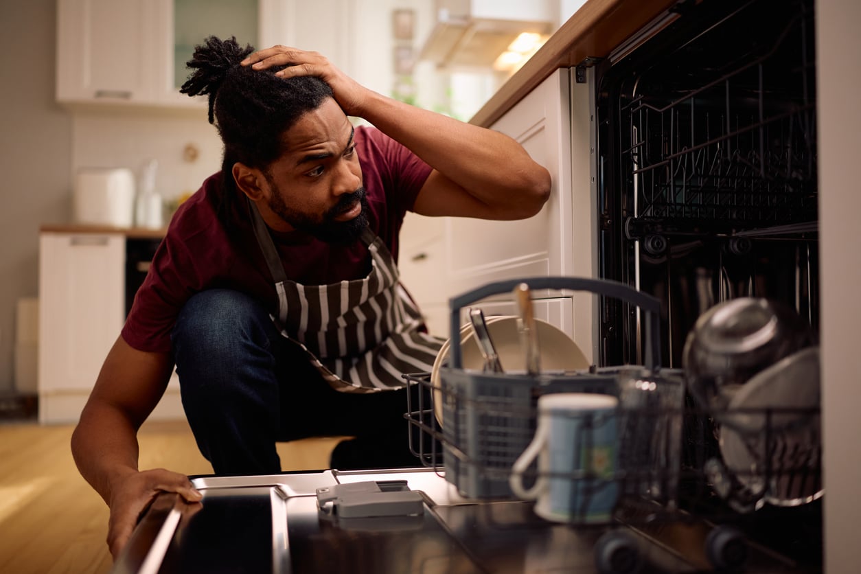 A man wearing an apron crouches in front of an open dishwasher, holding his head in confusion while inspecting the dishes inside a modern kitchen.
