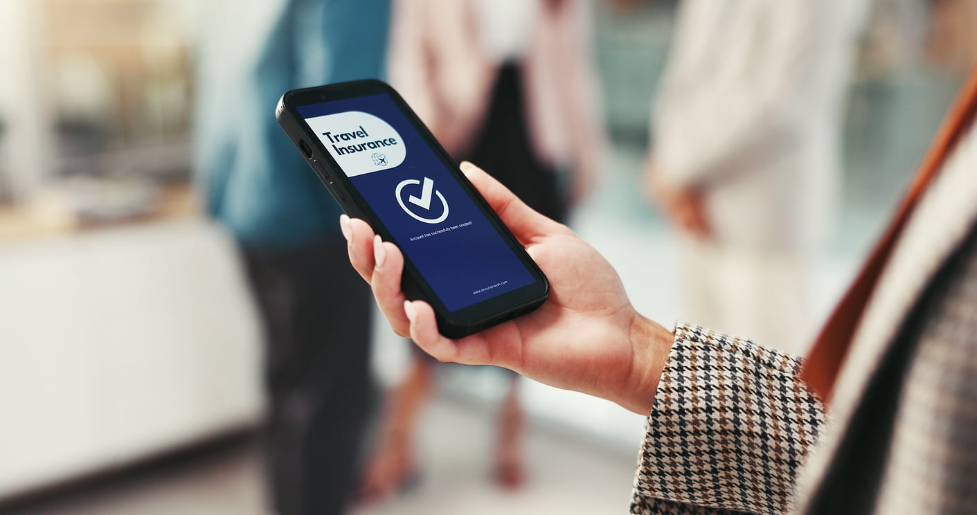 A person holds a smartphone displaying a travel insurance app with a check mark on the screen, confirming their coverage before booking a flight. The individual wears a plaid coat, while blurred people and a bright indoor setting appear in the background.