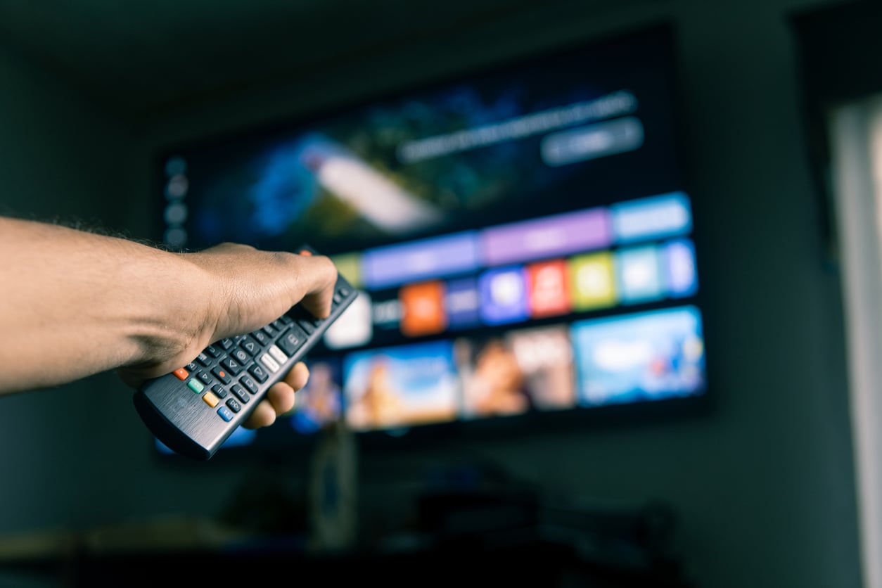 A hand holding a remote control points at a large TV screen displaying colorful apps and streaming options in a blurred living room, highlighting money saving habits by choosing affordable entertainment alternatives.