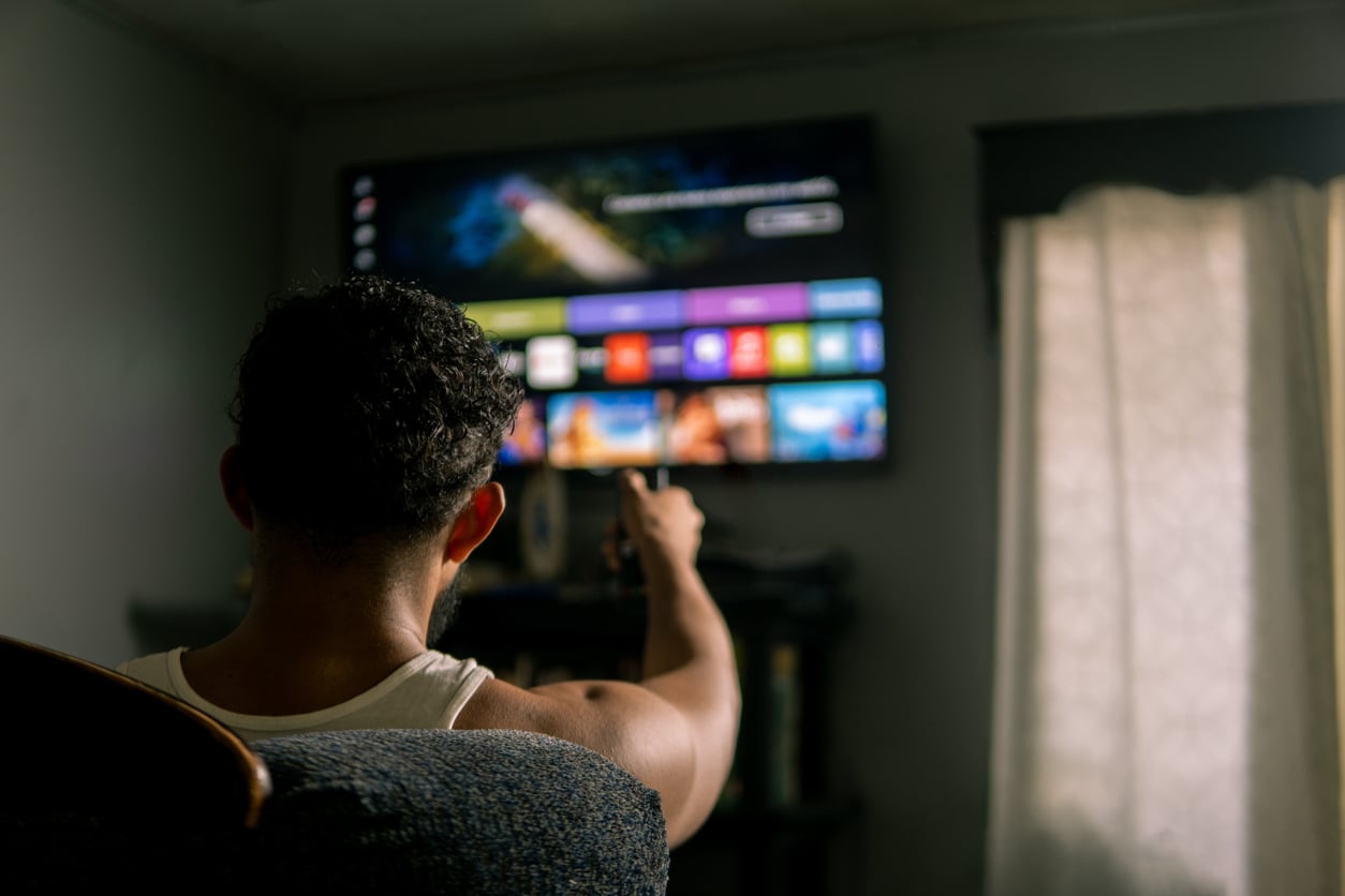 A person sits in a chair, pointing a remote at a large, wall-mounted smart TV displaying a colorful menu in a dimly lit room—an inviting setup for relaxing while tracking household expenses.