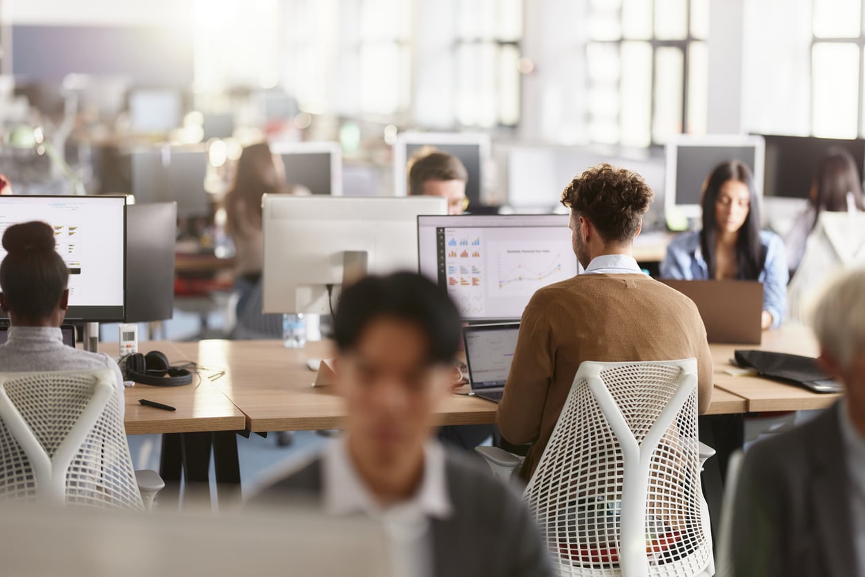 A bright, modern open-plan office where multiple employees sit at desks, working on computers displaying charts and graphs. The scene is viewed from behind, highlighting a collaborative, professional environment that values teamwork and productivity—principles often associated with traditional Baby Boomer beliefs—as colleagues remain focused on their screens.