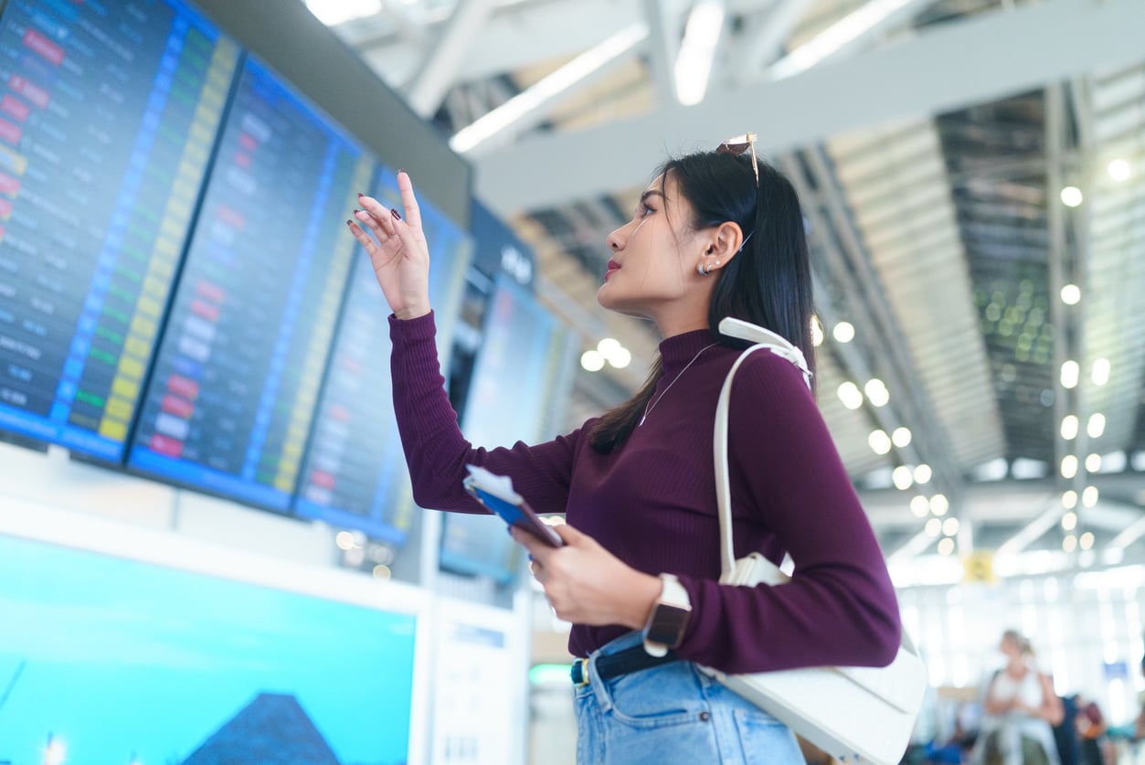 A woman in a purple top points at a digital flight information board in an airport, holding her phone and passport with her other hand, carefully checking details before booking a flight. Bright airport lights and a large board with flight details are visible.