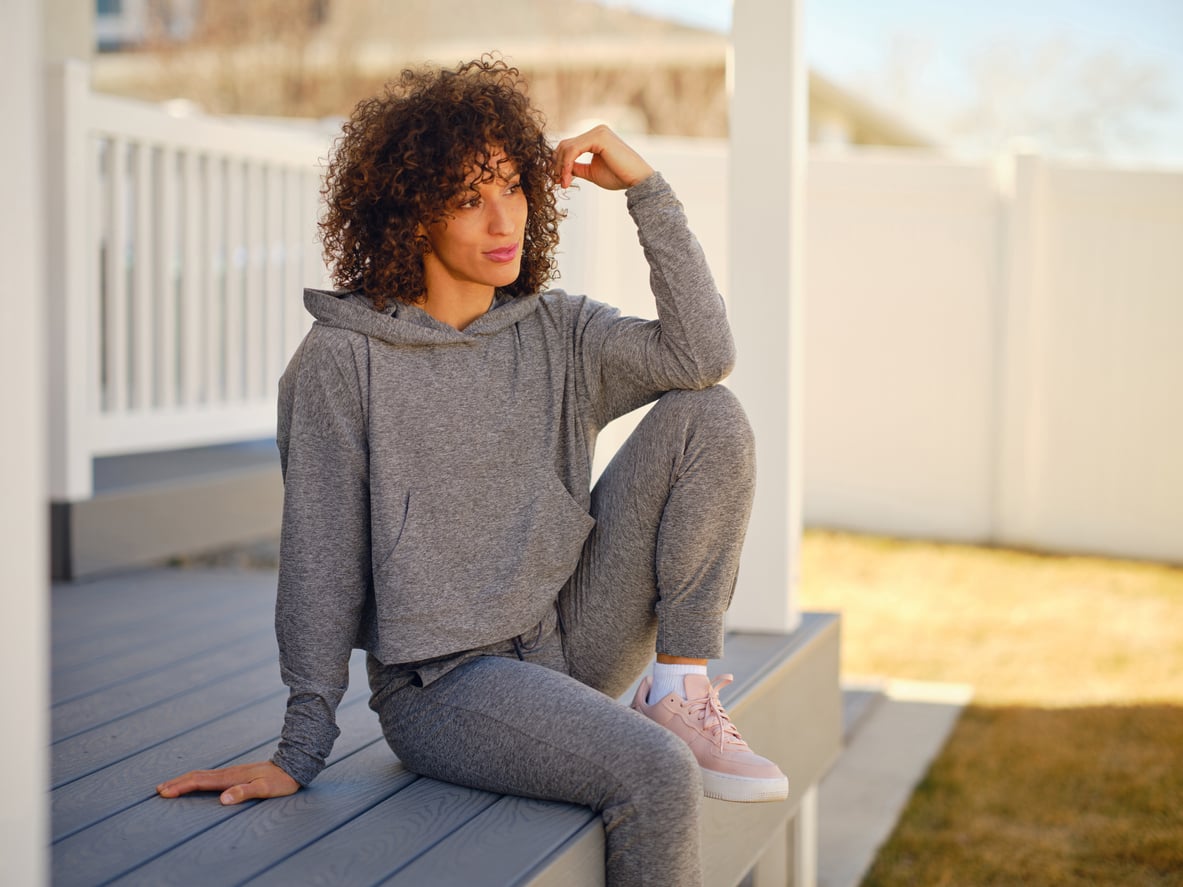 A person with curly hair in a gray hoodie and sweatpants sits on outdoor steps, resting one arm on their knee and looking into the distance.