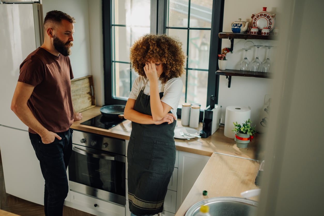 A woman in an apron stands in a kitchen, covering her face with her hand, while a man looks at her. The atmosphere appears tense or emotional.