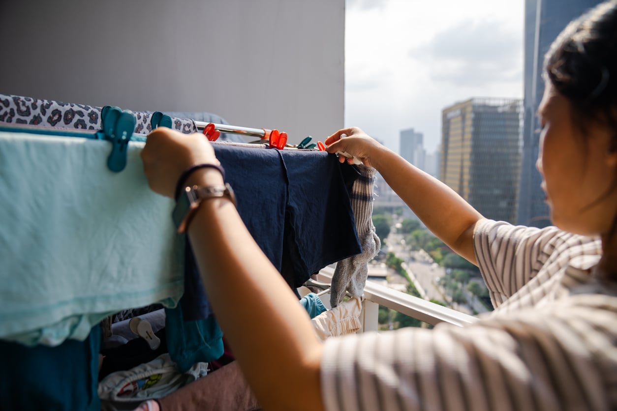 A person hangs clothes to dry on a balcony clothes rack, with tall buildings and a cityscape visible in the background on a sunny day.