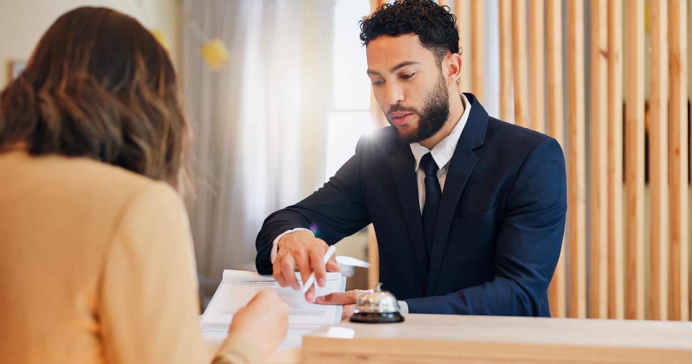 A man in a suit sits at a reception desk, pointing to a document while assisting a seated woman with her hotel booking mistakes. A service bell is on the desk in front of them.
