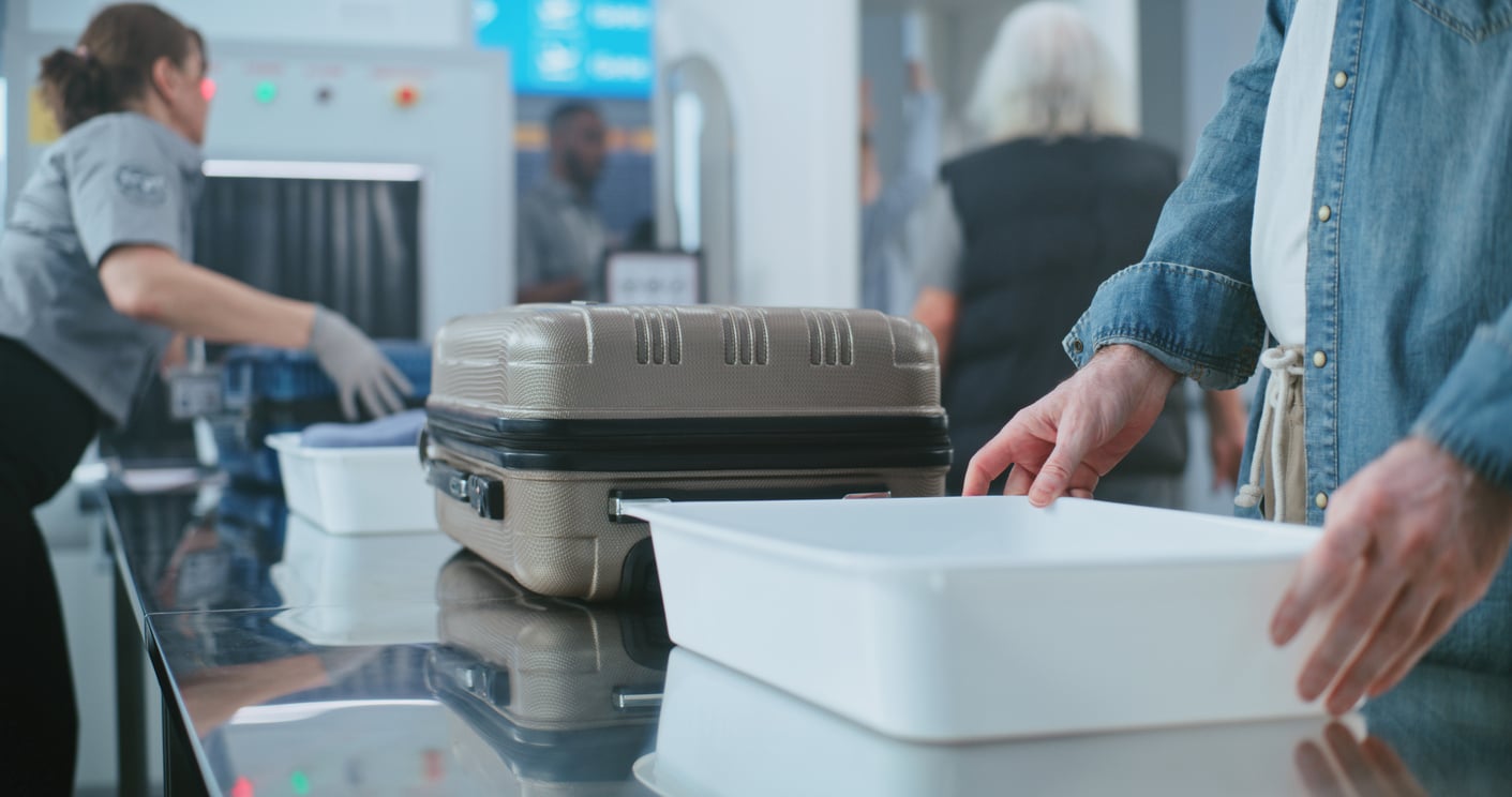 A person places a suitcase in a white tray at an airport security checkpoint, perhaps thinking about beach vacation savings, while security staff work in the background.