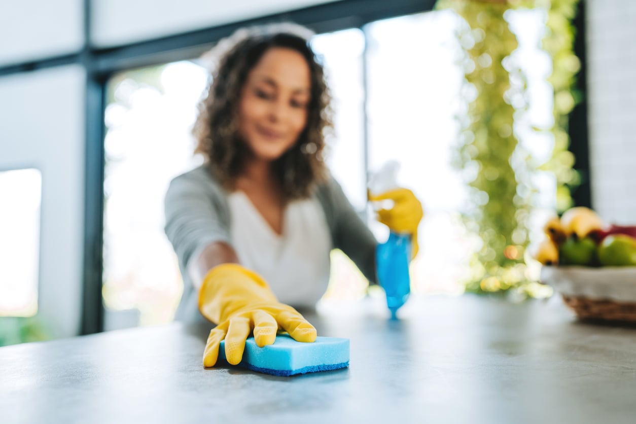 Woman wearing yellow gloves cleans a kitchen counter with a blue sponge, holding a spray bottle—once among the most popular products people no longer buy as frequently today. The background features bright windows and a fruit basket. The focus is on her hand and cleaning supplies.