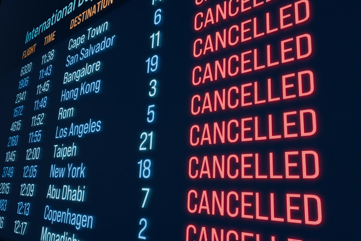 Airport departure board showing multiple international flights with red "CANCELLED" notices next to each destination, including Cape Town, Hong Kong, Los Angeles, and New York—a stark reminder to always check the latest travel updates before booking a flight.