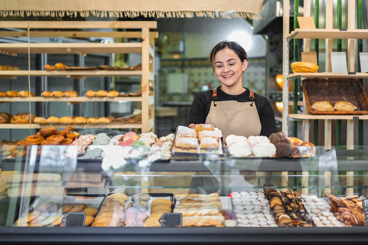 A smiling woman wearing an apron stands behind a glass display case filled with assorted pastries and baked goods in a bakery, perhaps sharing her recipes as part of vacation swaps. Shelves with more bread and pastries are in the background.