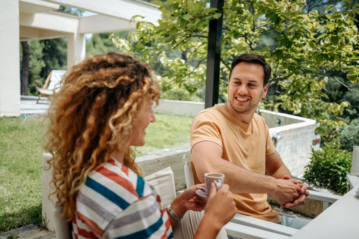 A woman and a man sit outside on a patio, smiling and talking. The woman holds a cup, and greenery surrounds them. With stylish outdoor home products enhancing the space, the setting appears relaxed and summery.