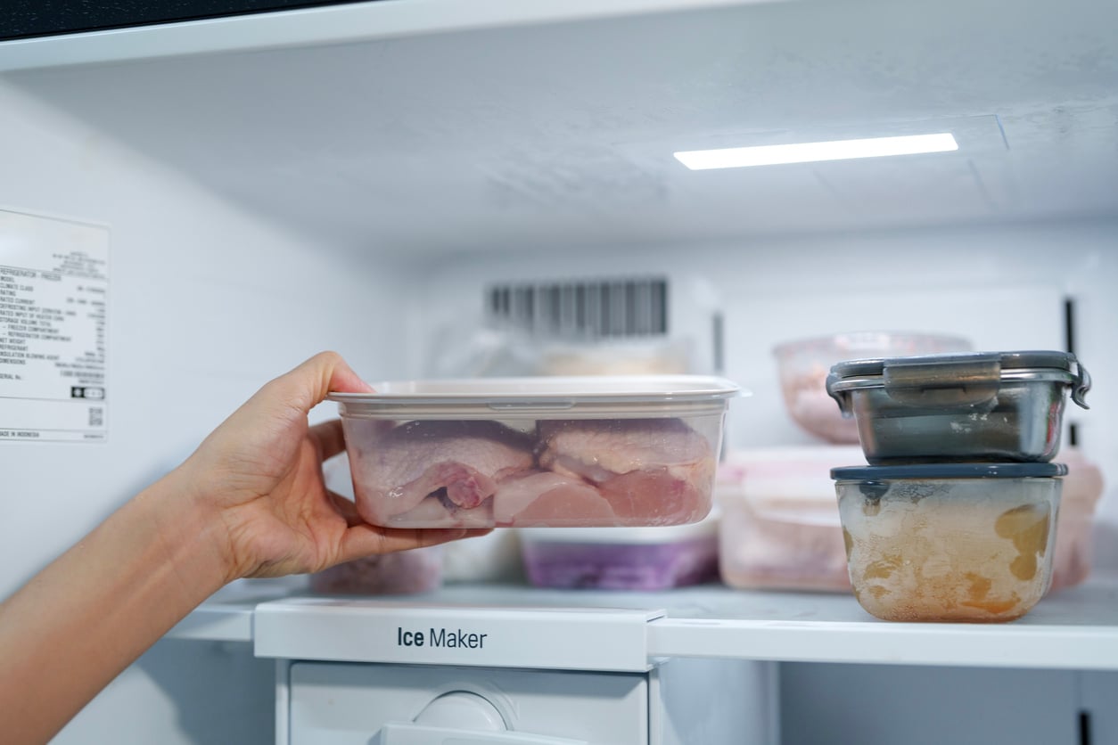 A hand places a plastic container of raw chicken pieces into a freezer, alongside other sealed containers filled with various frozen foods.