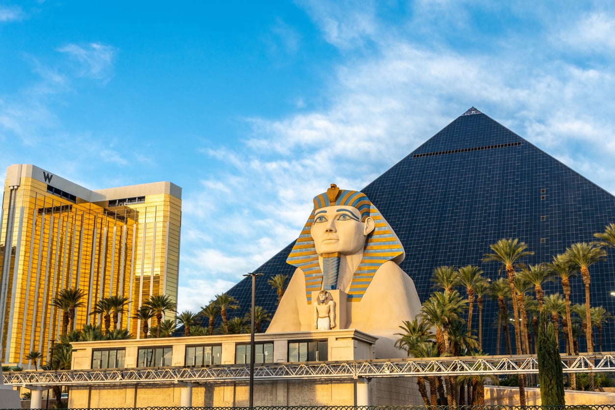 A large Sphinx replica and a glass pyramid hotel at the Luxor Las Vegas, with palm trees and a golden building nearby under a partly cloudy sky.