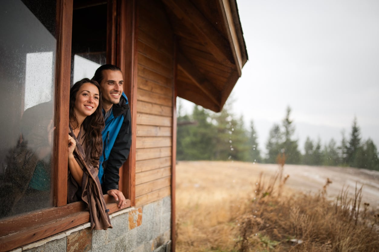 A smiling man and woman lean out of a wooden cabin window, looking outside. Despite the rain and misty mountains in the background, they seem carefree—perhaps enjoying a getaway where vacation costs are outweighed by spectacular views of the forest.