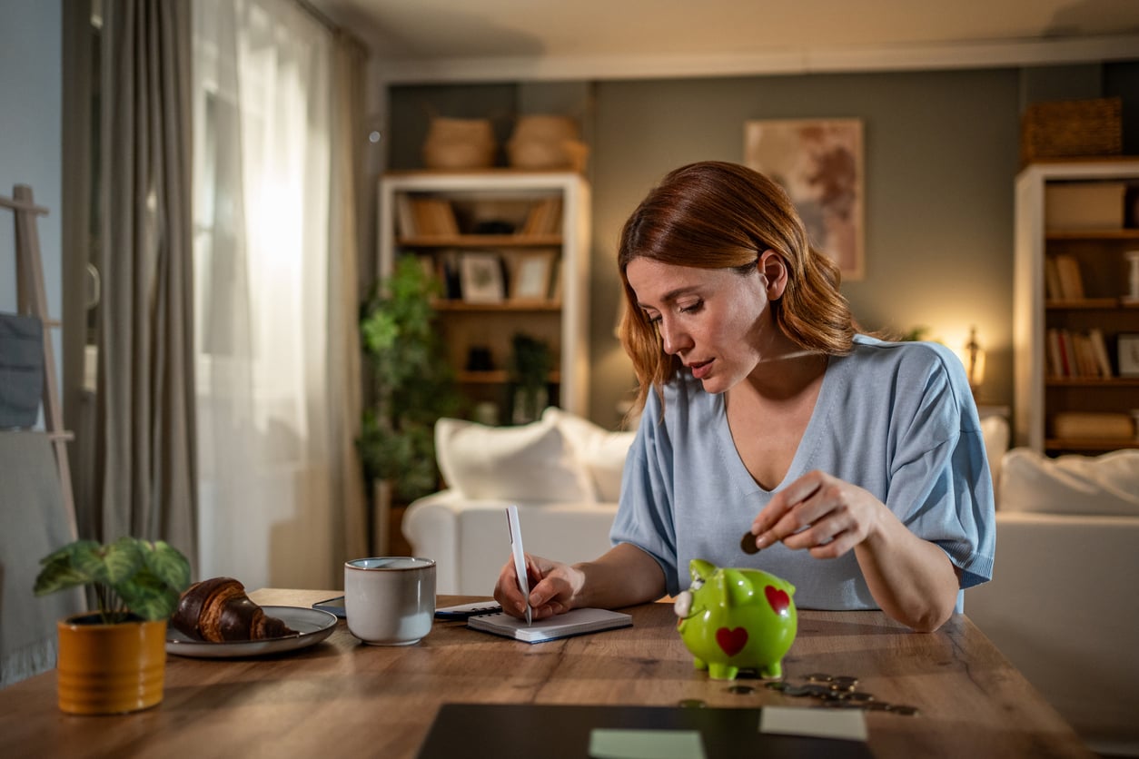 A woman sits at a table in a cozy living room, writing in a notebook while holding a coin over a green piggy bank. There is a coffee mug, croissant, and some coins on the table.