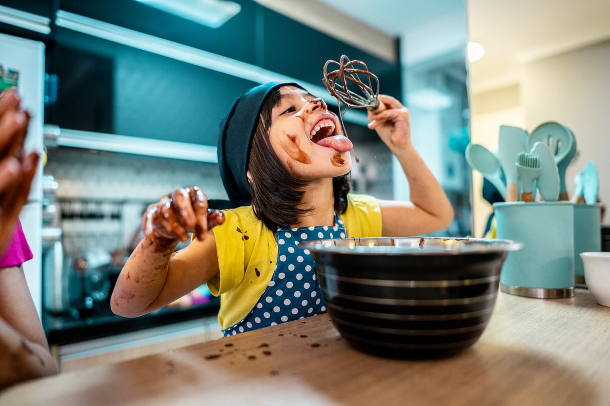 A young girl wearing a chef hat and polka-dot apron joyfully licks chocolate off a whisk, her face and hands messy, sitting at a kitchen table with a mixing bowl in front of her.