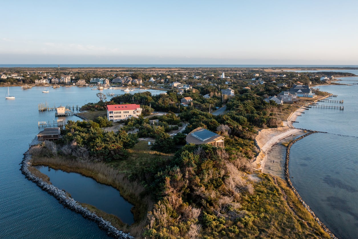 Aerial view of a coastal village with houses surrounded by trees, bordered by calm blue water and small beaches, under a clear sky—a picturesque scene reminiscent of charming beach towns.