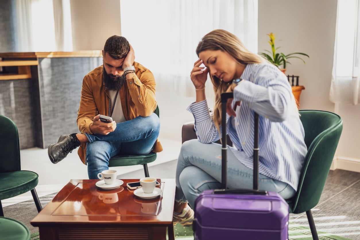 A man and woman look stressed while sitting in a waiting area with coffee cups and a purple suitcase, possibly experiencing travel delays due to hotel booking mistakes.