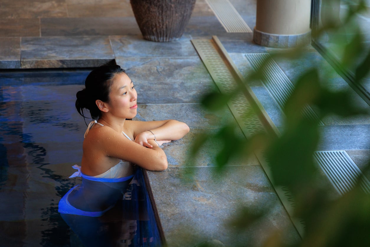 A woman in a white bikini relaxes in an indoor pool, leaning on the edge and looking thoughtfully toward a window, perhaps reflecting on past hotel booking mistakes. Green leaves are visible in the foreground.