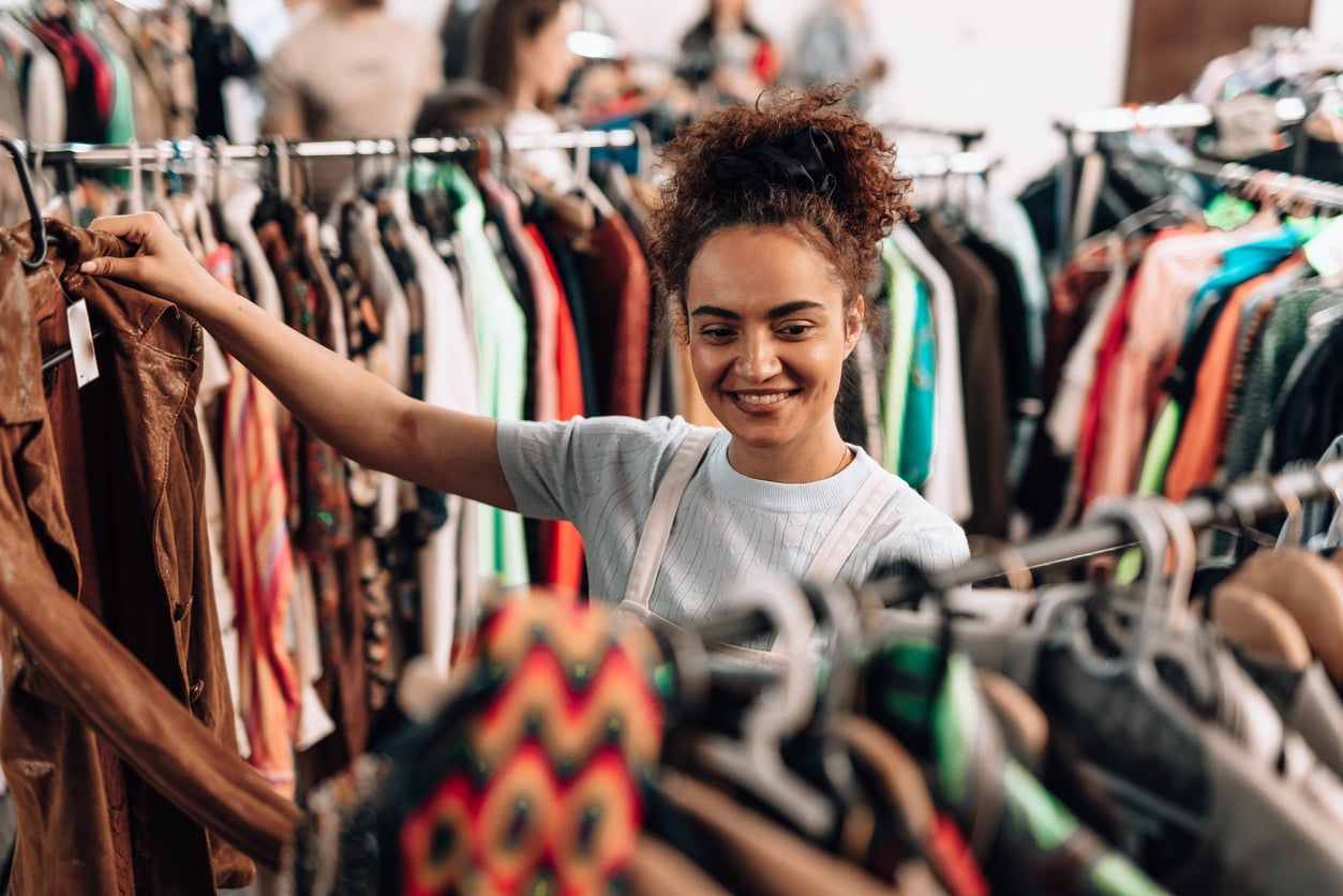 A woman with curly hair smiles while browsing clothes on a rack in a busy thrift store, reflecting how Women Are Changing Their Spending Behaviors. She is reaching for a brown jacket among various colorful garments.