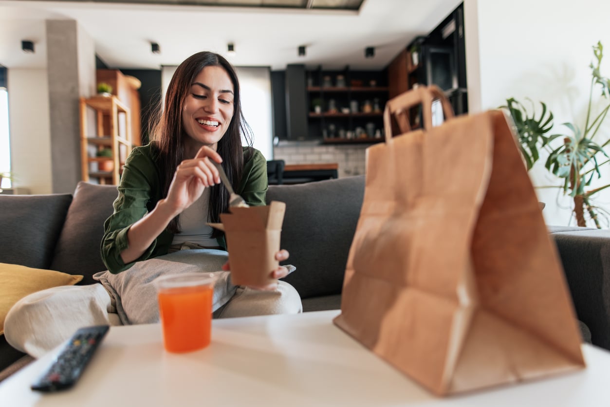 A woman sits on a couch, smiling while eating takeout food from a container. A drink, a remote, and a paper bag are on the white table in front of her. Carefree and relaxed, she enjoys her meal in a modern, stylish living room—momentarily setting aside thoughts of work or monthly bills.