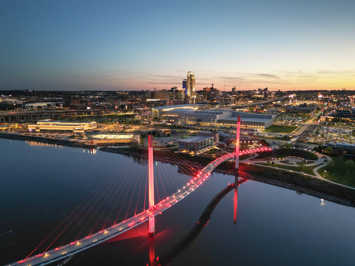 A travel-friendly, lit-up cable-stayed pedestrian bridge crosses a river at dusk, with a city skyline and illuminated buildings in the background.
