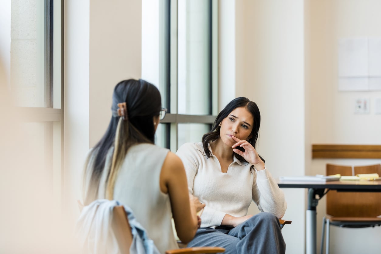 Two women sit in a bright room having a serious conversation about life before smartphones. One woman listens attentively with her hand on her chin, reflecting on the simplicity of past connections, while the other speaks, her back facing the camera.