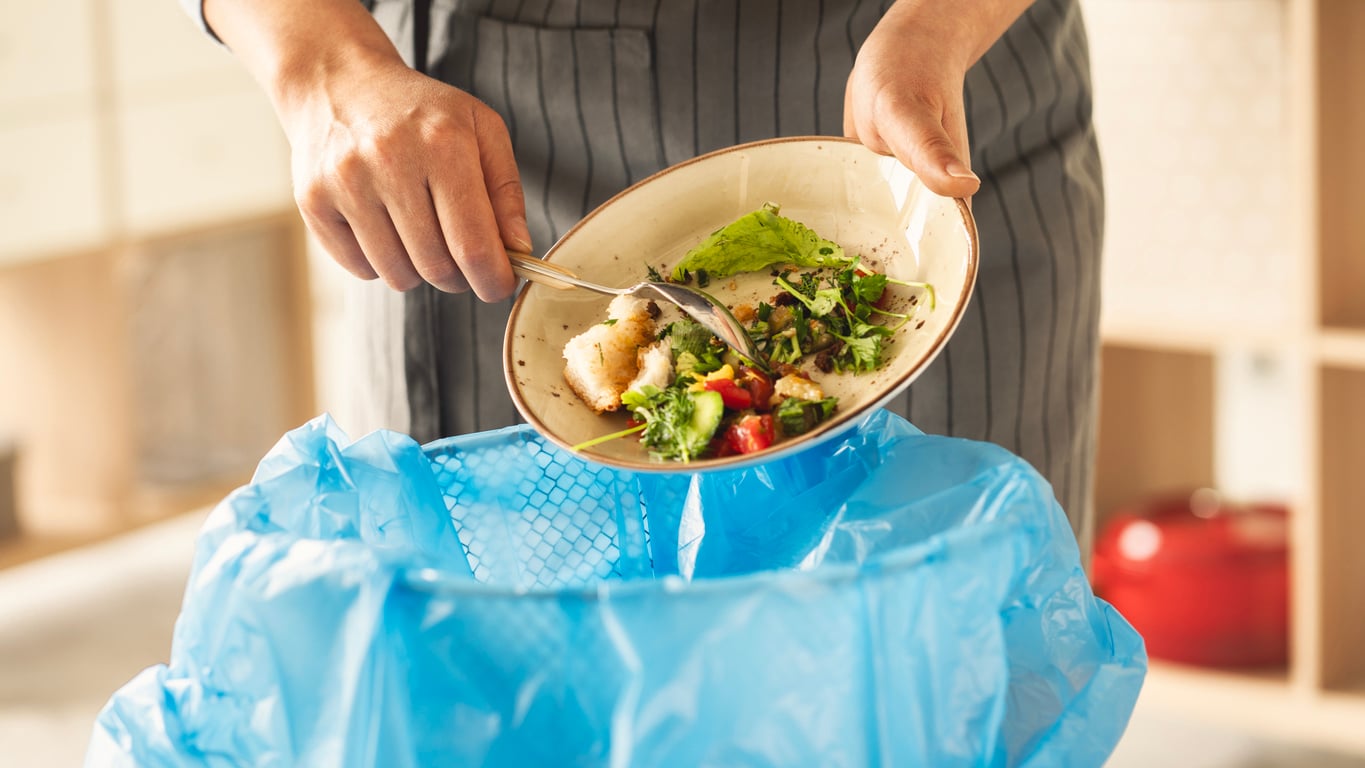 A person scraping leftover food from a plate into a trash bin lined with a blue plastic bag, helping to reduce household expenses by minimizing food waste.