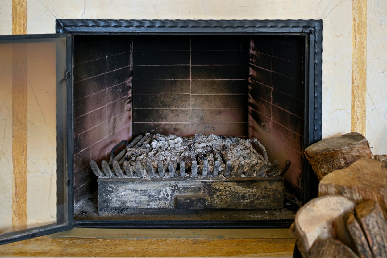 An open glass-door fireplace with a metal grate holding gray ashes reflects one of the home features homeowners no longer want. Stacked firewood is visible on the right side of the image, while the fireplace interior is blackened from previous fires.