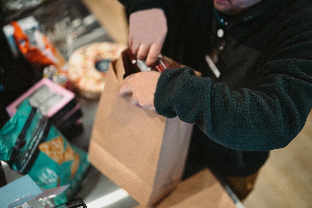 A person places groceries, including chips and snacks, into a brown paper bag at a checkout counter.