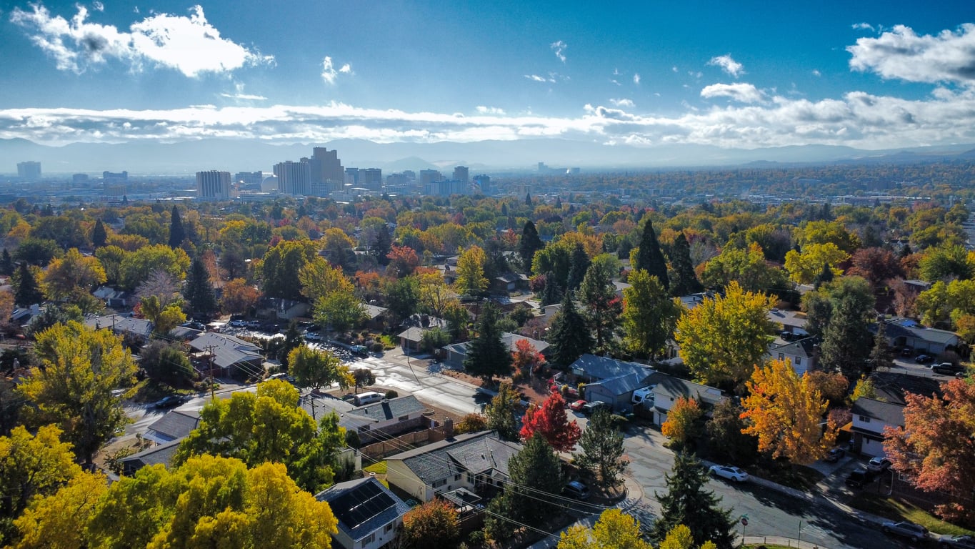Aerial view of a city with autumn trees and colorful foliage, suburban houses in the foreground, and tall buildings and distant mountains under a partly cloudy blue sky—an inviting scene that highlights one of the most affordable travel destinations for those seeking both natural beauty and urban charm.