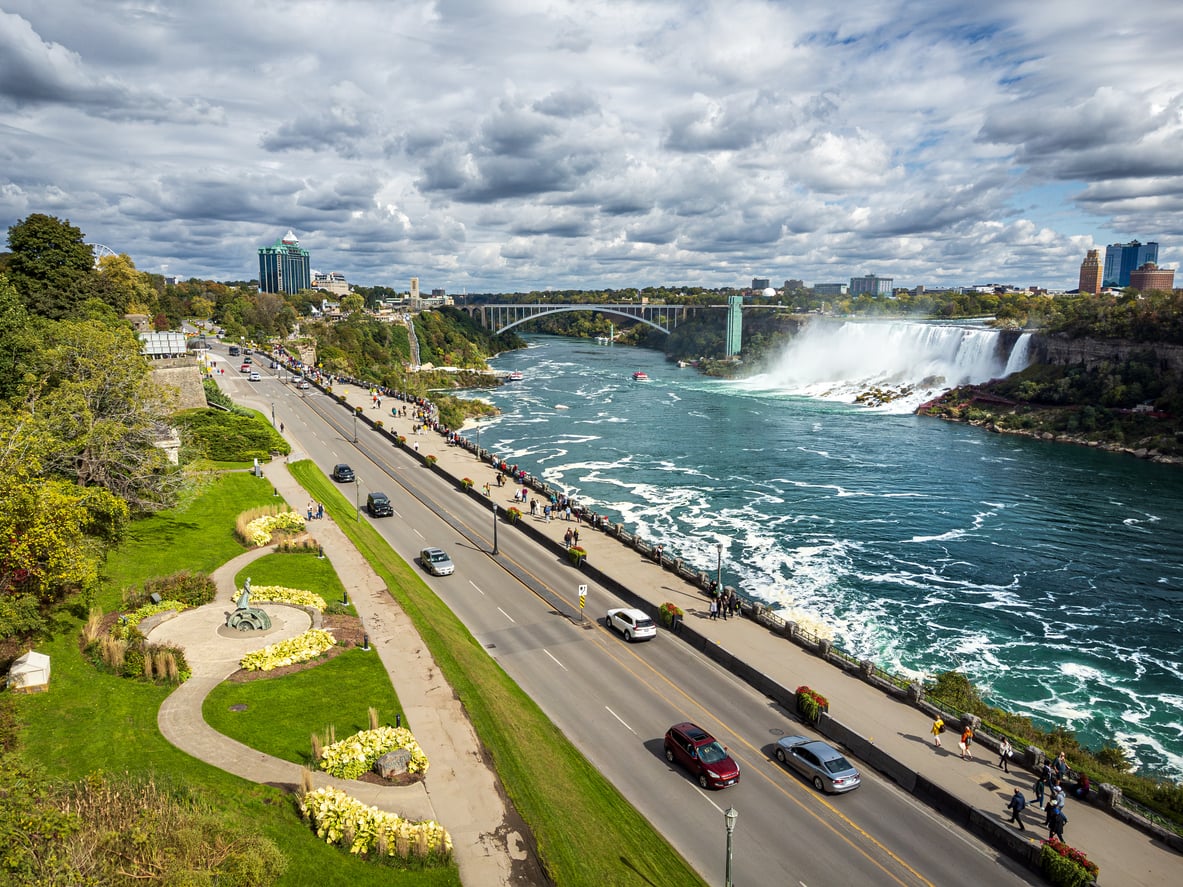 A scenic view of Niagara Falls—one of the world’s top 20 travel destinations—with cars on a road beside the river, people walking on pathways, green trees, gardens, and a distant bridge under a partly cloudy sky.