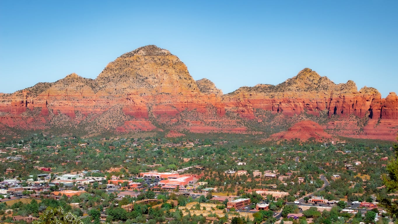 Red rock mountains rise behind a town with green trees and buildings under a clear blue sky, creating a scenic desert landscape that stands out among top travel destinations.