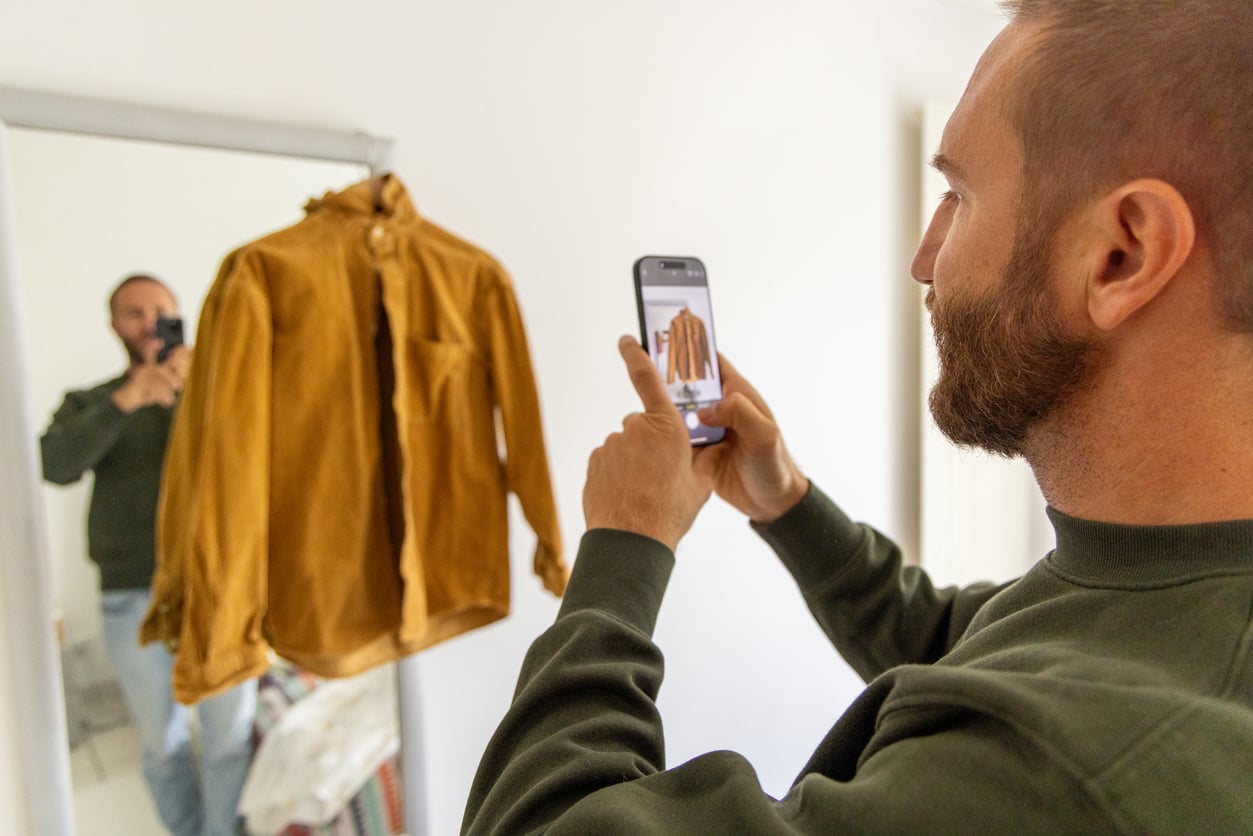 A man uses his smartphone to take a photo of a brown jacket hanging on a mirror, with his reflection visible in the background.