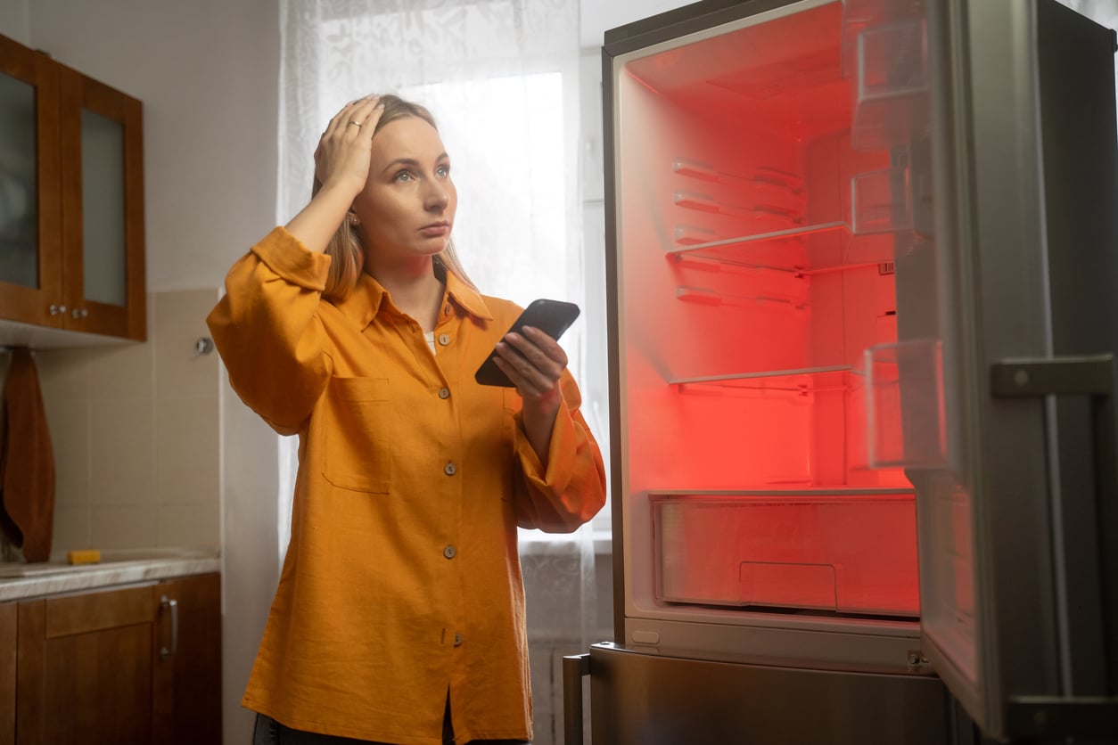A woman in an orange shirt stands in a kitchen, holding a phone and looking worriedly at an open refrigerator with a red light inside, frustrated by products that don't last.