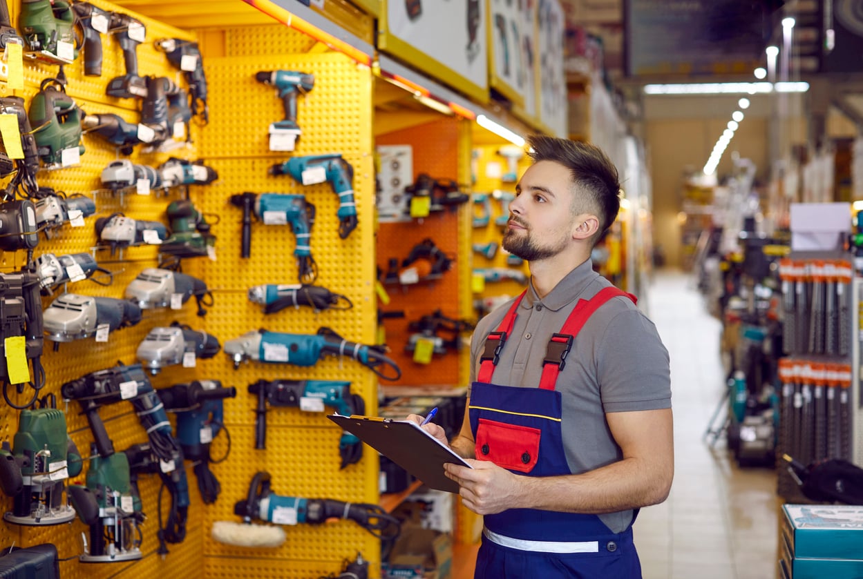 A hardware store employee in overalls holds a clipboard and inspects power tools displayed on a wall in a brightly lit store aisle.