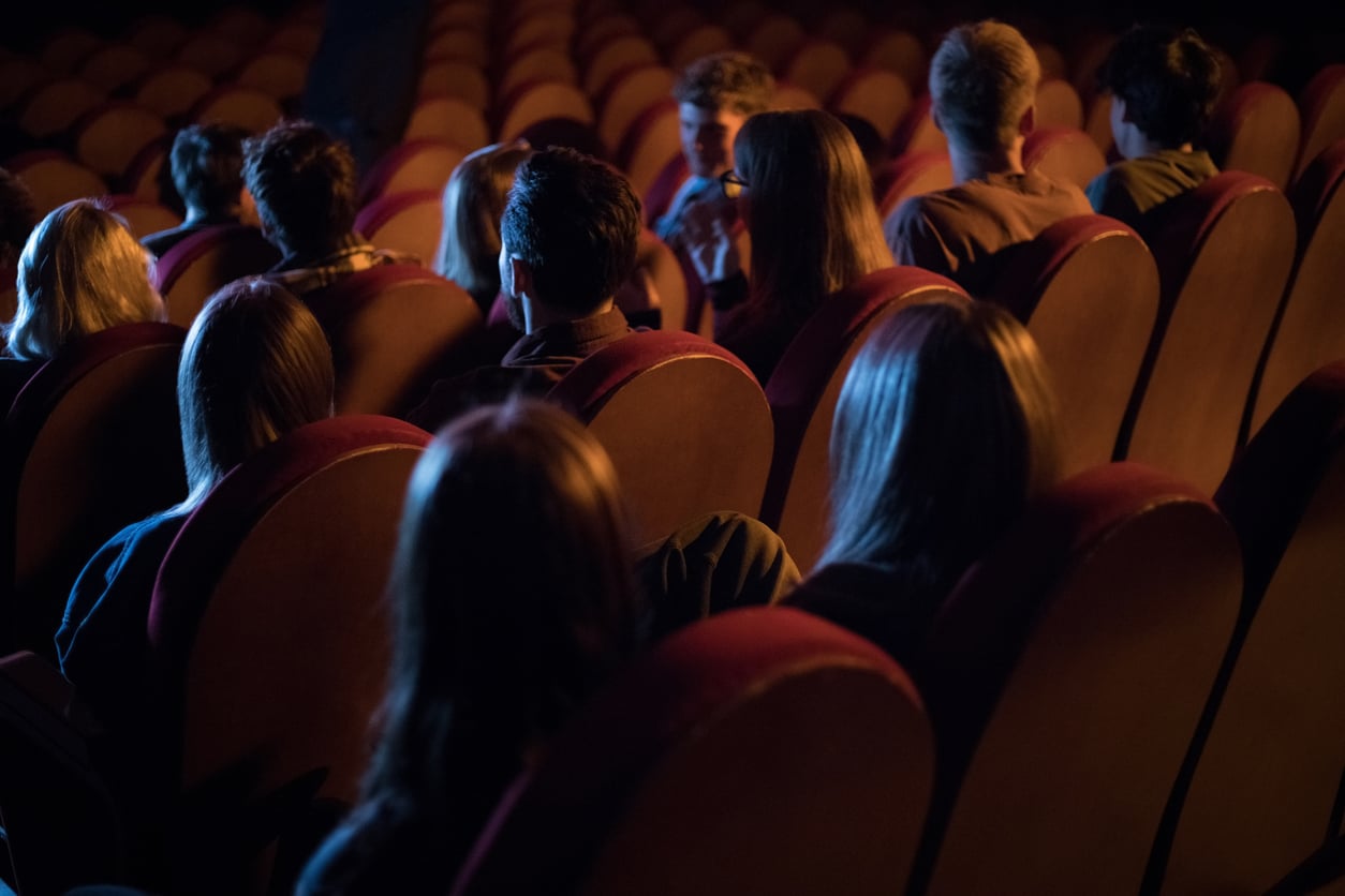 A dimly lit theater shows people sitting in rows of red-cushioned seats, many unoccupied—perfect for trying out a few discreet movie theater hacks while enjoying the show.