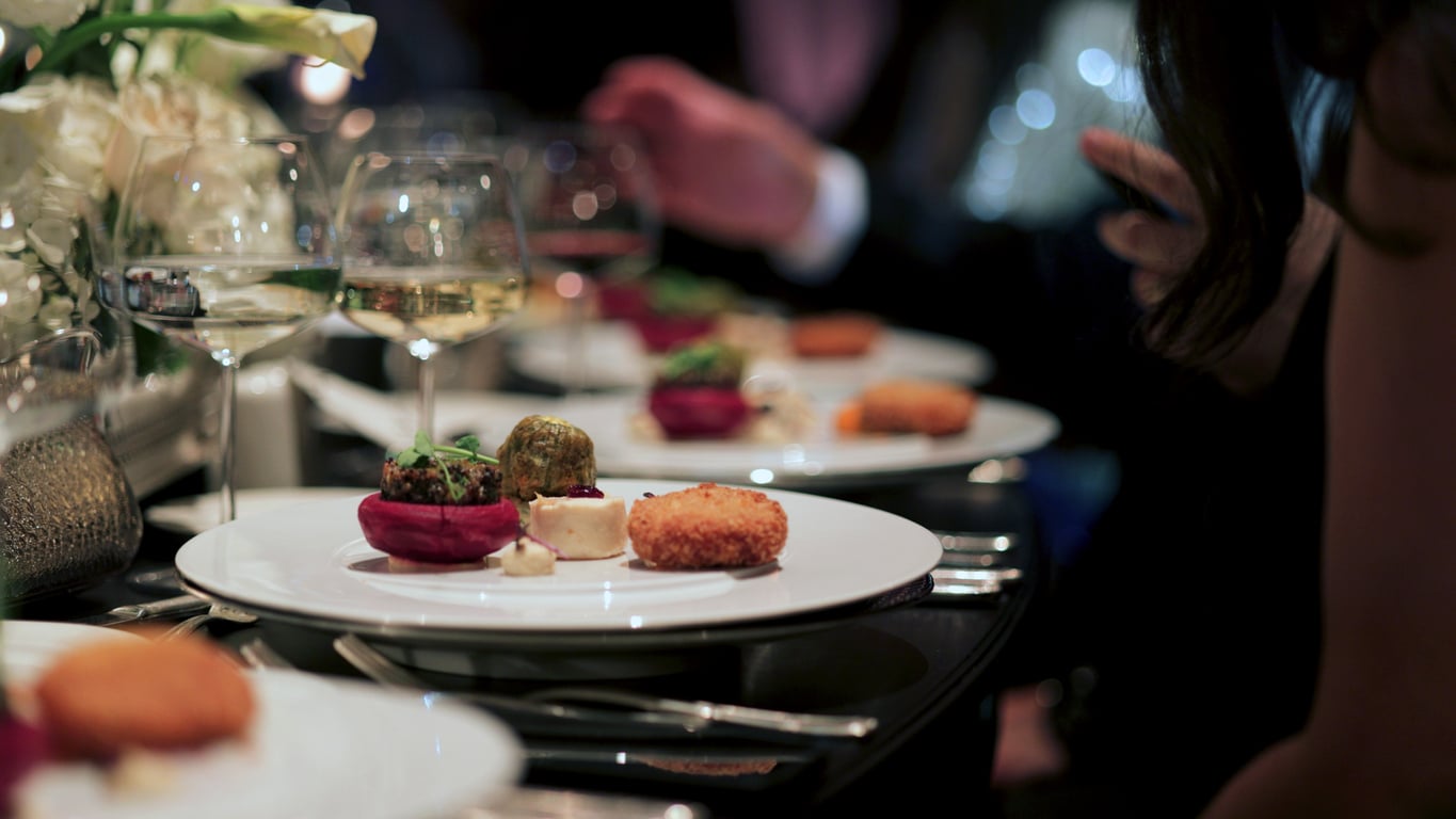 A close-up of elegant dinner plates with gourmet appetizers, wine glasses, and people dressed formally, sitting at a stylishly set dining table.