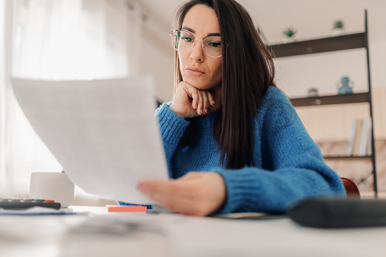 A woman wearing glasses and a blue sweater sits at a desk, looking thoughtfully at a document she is holding—one of her monthly bills. A shelf and window are visible in the background.
