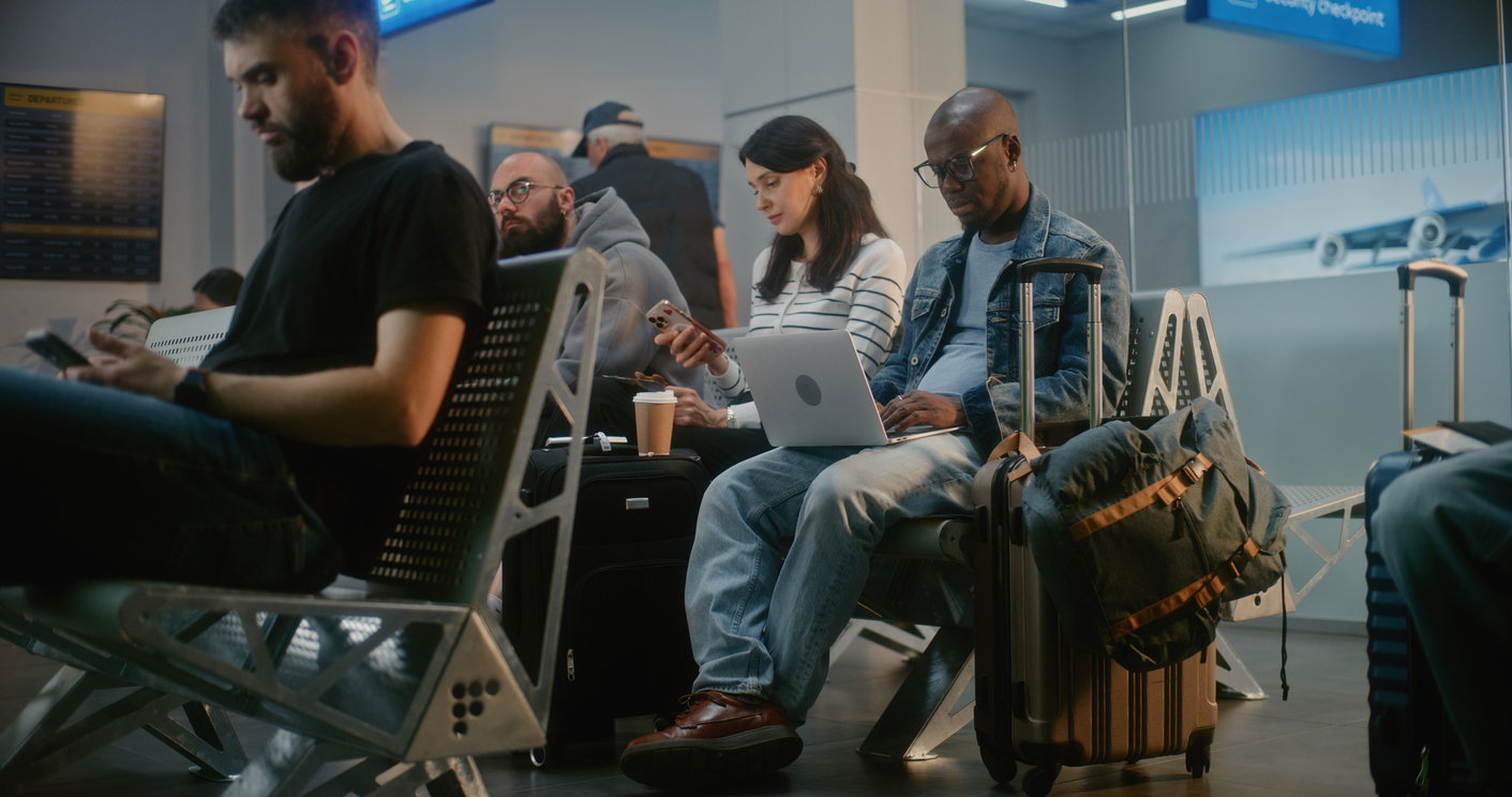 People sit in an airport waiting area with luggage, using laptops and phones. Departure screens are visible above, and a large window shows part of an airplane outside.